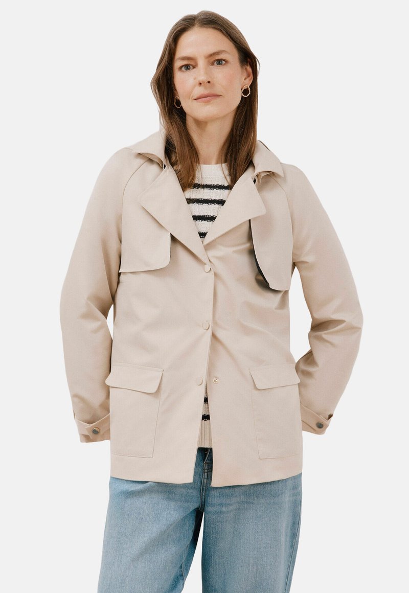 Beige, knäppt jacka med krage, två framfickor och ett lager av bröstfickan. Bärs över en randig tröja och ljusblå jeans.