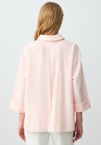 Camicia a maniche lunghe rosa chiaro con vestibilità rilassata, colletto e polsini arrotolati. Tessuto textura con cucitura centrale e spalle scese.