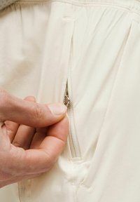 Bežinės kelnės su šoniniu užtrauktuku. Rankos sukandusi užtrauktuko kilpą, demonstruojantis sklandų audinio paviršių ir elastingą juostos detalę.