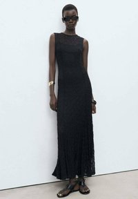 Robe noire sans manches en dentelle avec un bas évasé, présentant un motif texturé. Les accessoires comprennent un bracelet en or et des sandales noires.