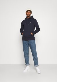 Sudadera azul marino con cordón, bolsillo tipo canguro y logotipo bordado. Combinada con jeans azules remangados y zapatillas blancas.