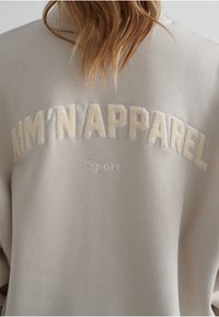 Světle béžová mikina s vyvýšeným krémovým vyšitým textem "AIM'N'APPAREL" a "Sport" na zadní straně. Měkčená textura, ležérní design.