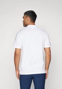Polo bianco con colletto a due bottoni e righe laterali blu. Il tessuto sembra liscio, con maniche corte e fondo orlato.