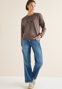 Sudadera marrón con estampado "JOYFUL", combinada con vaqueros acampanados azul claro y zapatillas beige. Textura suave, ajuste casual y mangas largas.