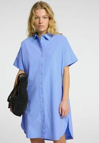 Abito blu chiaro con bottoni, maniche corte, colletto e spacchi laterali. Presenta una vestibilità ampia e un design minimalista. Accessorio: borsa nera intrecciata.