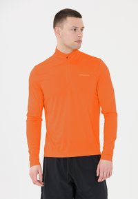 Oranssi pitkähihainen urheilupaita, jossa on neljänneksen vetoketju kauluksessa, valmistettu kevyestä teksturoidusta kankaasta. Rinnassa logo.