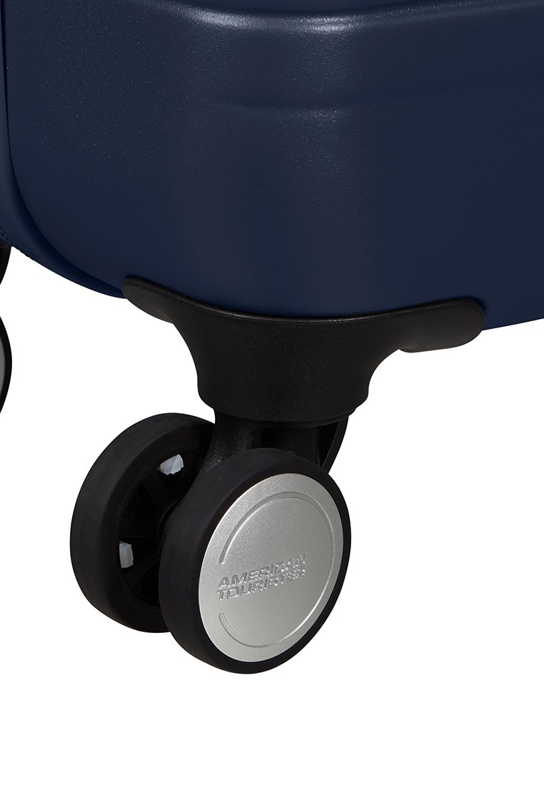 Angolo di una valigia rigida navy con attacco per ruote in plastica nera e mozzo della ruota argento con logo "American Tourister" in rilievo.