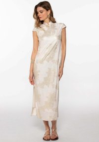 Robe beige clair avec un col haut, des manches courtes et un tissu brillant texturé présentant des motifs abstraits dorés ; fente sur le côté et sandales plates.