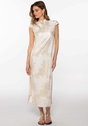 Vestido beige claro con cuello alto, mangas cortas y tejido brillante texturizado que presenta patrones abstractos dorados; abertura lateral y sandalias planas.