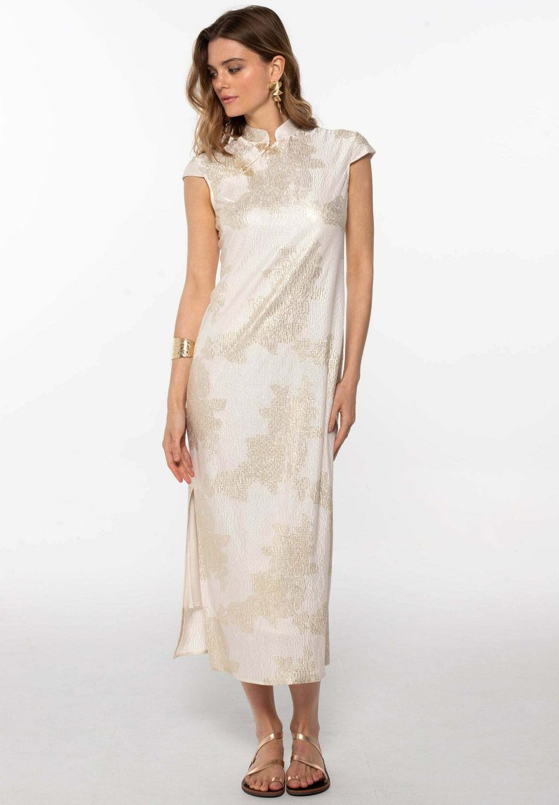 Robe beige clair avec un col haut, des manches courtes et un tissu brillant texturé présentant des motifs abstraits dorés ; fente sur le côté et sandales plates.
