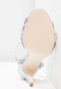 Sandał na wysokim obcasie z gładką, srebrną cholewką, lawendowymi akcentami z materiału i spiczastym noskiem. Podeszwa jest jasnobeżowa z teksturą o żłobieniach.