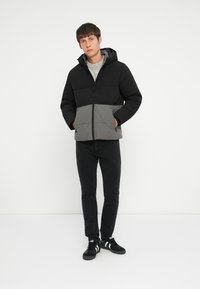 Polstret jakke med en sort top og grå midtersektion, med hætte, frontlynlås og sidelommer. Bæres over en grå sweater.