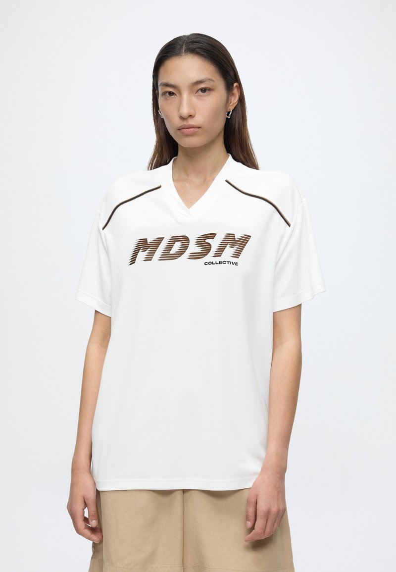 Jeune femme aux longs cheveux portant un t-shirt blanc MDSN Collective et un short beige, debout devant un fond clair uni.