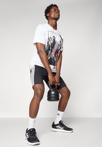 Hvit grafisk t-skjorte med svart og rødt design, svarte shorts med hvite striper, svart kettlebell og svarte treningssko.