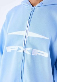 Sudadera con cremallera de color azul claro, hecha de tela suave. Presenta un diseño de logo en blanco "PXP" en el pecho y una capucha con cordón. Textura suave.