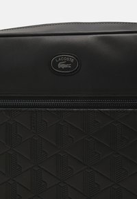 Svart Lacoste-veske med en glatt tekstur og et preget geometrisk mønster. Inkluderer en glidelås-lukking og et merket logo-patch.