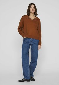 Suéter marrón de punto con cuello, combinado con jeans de mezclilla azules. El suéter tiene un corte holgado y puños acanalados; los zapatos son mocasines negros y brillantes.