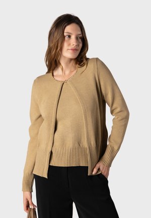 Femme portant un pull en maille beige et un cardigan assorti avec un pantalon noir, tenant un sac à main beige dans une main.
