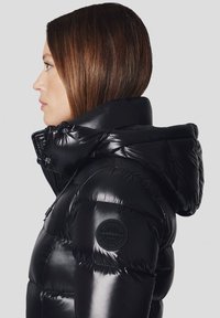 Czarna kurtka puchowa o błyszczącym wykończeniu, z kapturem i przeszyciami. Posiada okrągłą naszywkę z logo na lewym rękawie. Zapinana na zamek z przodu.