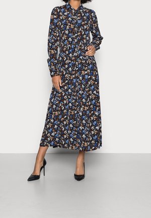 Femme portant une robe noire à manches longues boutonnée avec des motifs floraux bleus, blancs et bruns et des talons hauts pointus noirs.
