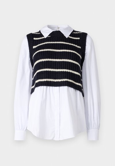 Stickad marinblå väst med vita ränder över en vit krag- och knapp-up skjorta. Vesten har en kortad design och texturerad tyg.