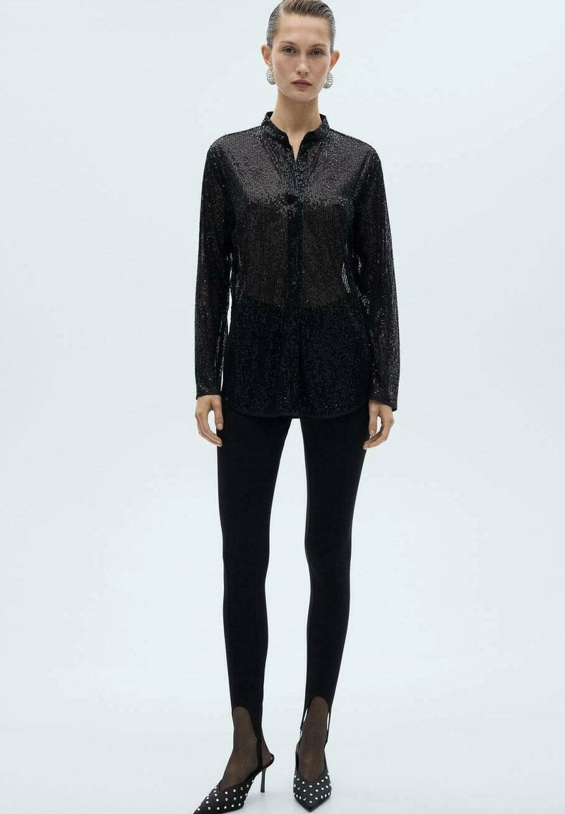 Sort pailletbluse med knaplukning, lange ærmer og rundet kant, sat sammen med sorte leggings og prikkede højhælede sko.