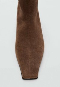 Bota de ante marrón con puntera cuadrada y puntiaguda, y textura suave y sin costuras sobre un fondo gris claro.