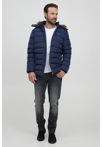 Marineblå pufferjakke med hette med pelskant, glidelås og sidelommer, kombinert med grå jeans og svarte støvler.