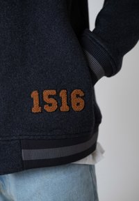 Dark navy jacket with brown tufted "1516" patch above ribbed gray-striped hem, worn with light blue jeans and hand in pocket.