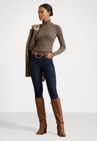 Bruine geribbelde coltrui, donkere denim jeans en kniehoge bruine leren laarzen met een blokhak. Bruine riem met kettingdetail.