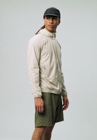 Veste beige légère avec fermeture éclair, manches longues et poignets élastiques, associée à un short vert olive, sur fond uni.