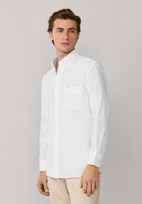 Giovane uomo che indossa una camicia bianca a maniche lunghe con bottoni e pantaloni beige, in piedi davanti a uno sfondo grigio chiaro, che guarda di lato.