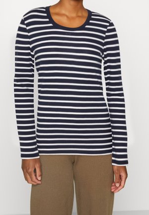 Personne portant un t-shirt à manches longues à rayures horizontales bleu marine et blanc et un pantalon marron, debout devant un fond uni.
