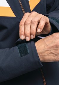 Marineblaues Jacket mit Reißverschluss und orangefarbenen Akzenten. Verfügt über eine Klettverschluss-Verstellung an den Ärmeln. Glattes, strukturiertes Material, Nahaufnahme einer Hand, die den Ärmel anpasst.
