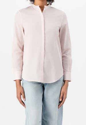 Femme portant une chemise rose clair à manches longues avec un col mandarin et un jean bleu clair, debout devant un fond blanc.
