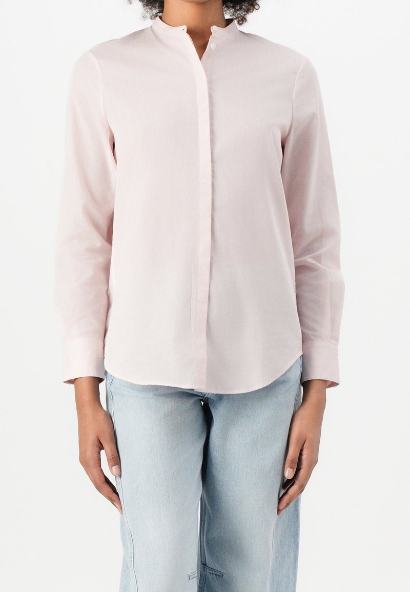 Femme portant une chemise rose clair à manches longues avec un col mandarin et un jean bleu clair, debout devant un fond blanc.