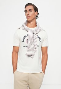 Junger Mann mit kurzen braunen Haaren, der ein weißes T-Shirt mit Aufdruck, beiges Hosen und ein gestreiftes Hemd über den Schultern trägt, mit den Händen in den Taschen.