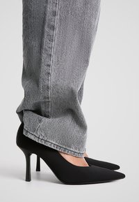 Svarta högklackade pumps med spetsig tå, gjorda av slätt material, i kombination med grå denimjeans som visar detalj av ankel och häl.