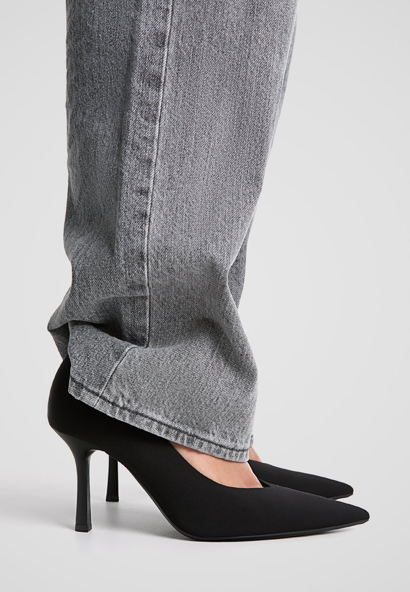Svarta högklackade pumps med spetsig tå, gjorda av slätt material, i kombination med grå denimjeans som visar detalj av ankel och häl.