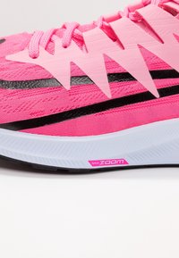 Zapatilla rosa con parte superior de malla, acentos negros y una superposición texturizada en rosa. Presenta la marca Nike Zoom en la entresuela blanca.