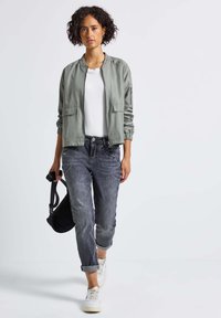 Veste bomber gris clair avec des poignets élastiques, associée à un jean gris délavé et des baskets blanches. Accessoire : petit sac à bandoulière noir.