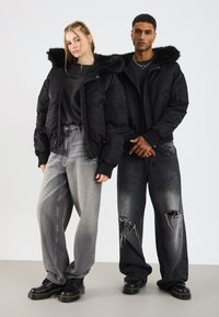 To modeller iført sorte jakker med hætte af pels, løse, falmede jeans og sorte støvler, står side om side mod en ensfarvet baggrund.