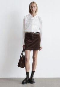 Woman wearing a white embroidered blouse, brown corduroy mini skirt with belt, dark socks, loafers, and holding a brown handbag.