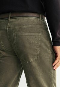 Olivgrüne Cordhosen mit strukturiertem Rippmuster, fünf Taschen und einem braunen Ledergürtel. Rückansicht zeigt das Taschendesign.