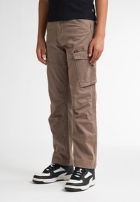 Bruine cargo-broek met meerdere zakken, rechte pasvorm, lichtgewicht stof en gestikte details. Draagt met zwart-witte sportschoenen.