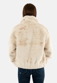 Giacca in pelliccia sintetica color crema, con colletto alto e elastico in vita, presenta una consistenza morbida e pelosa con sottili variazioni di tonalità.