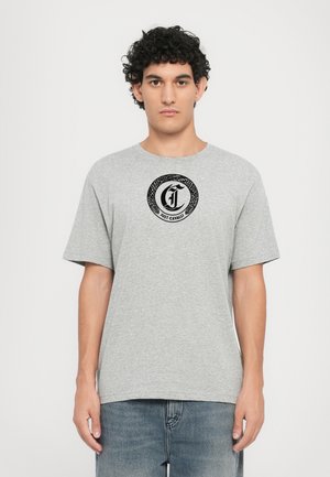Camiseta de algodón gris con cuello redondo y mangas cortas, que presenta un logo circular negro con texto en el centro frontal. Llevada con jeans azules.