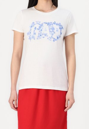 Vrouw draagt een wit T-shirt met korte mouwen met blauw bloemen "GAP" logo en een rode rok tegen een effen witte achtergrond.
