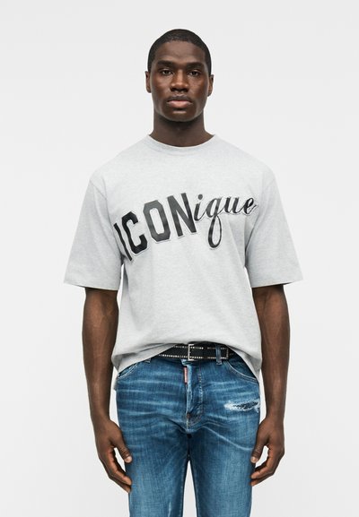 Νεαρός άνδρας που φοράει ανοιχτό γκρι μπλουζάκι με το κείμενο "ICONique" και μπλε τζιν με φθορές, στέκεται μπροστά σε απλό λευκό φόντο.