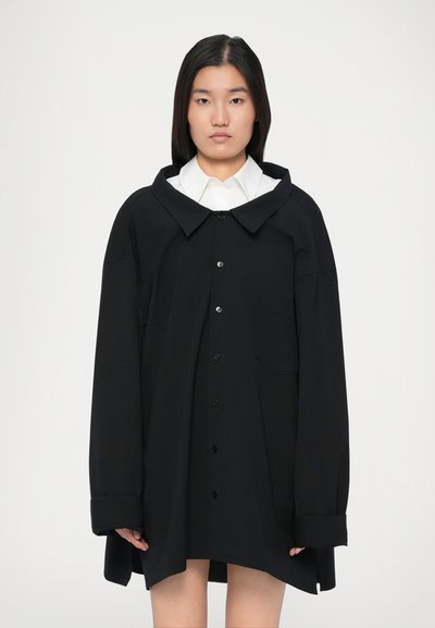 Tânără cu părul drept și închis la culoare, purtând un palton negru oversized cu nasturi peste o cămașă albă cu guler, stand pe un fundal simplu.